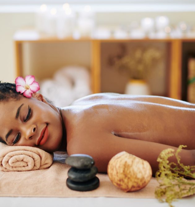 Beautiful young Black woman with flower in her hair resting on bed in spa salon after relaxing back massage with oils